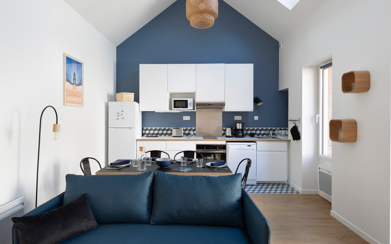 Cuisine blanche, salle à manger avec une table et chaises noires et le salon avec un canapé bleu.