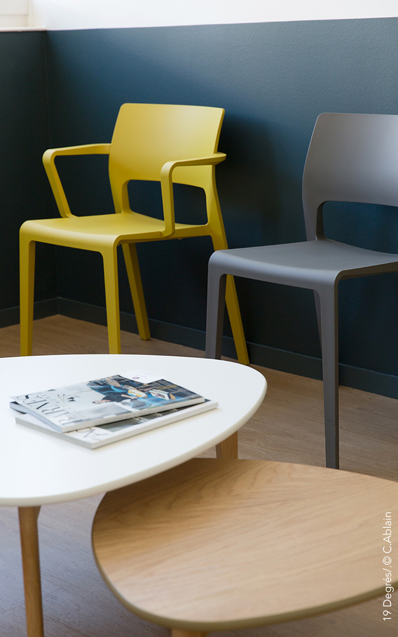 Salle d'attente du cabinet de neurologie avec des chaises en plastique dur grises et jaunes ainsi que des tables gigognes.