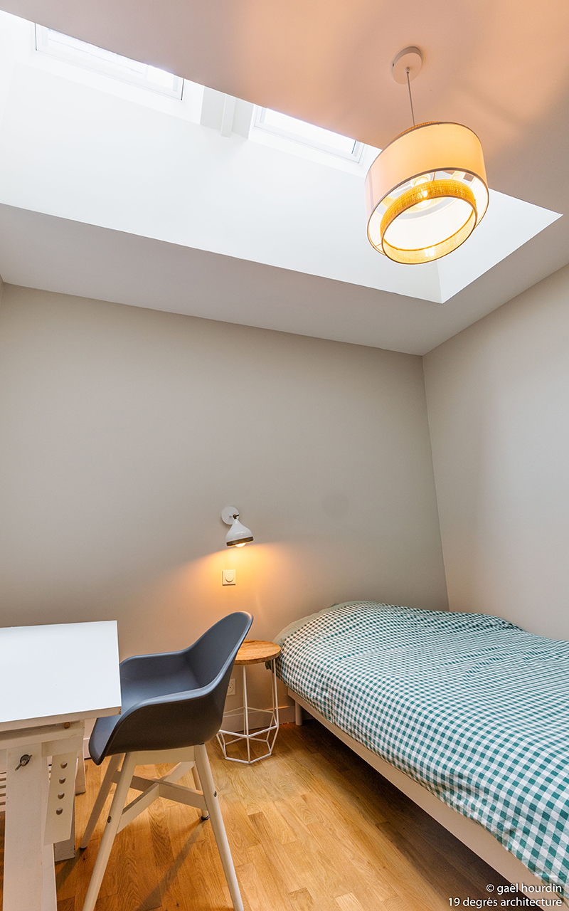 Chambre simple avec un lit, une table de chevet et sa lumière ainsi qu'un bureau blanc et une chaise grise et blanche.