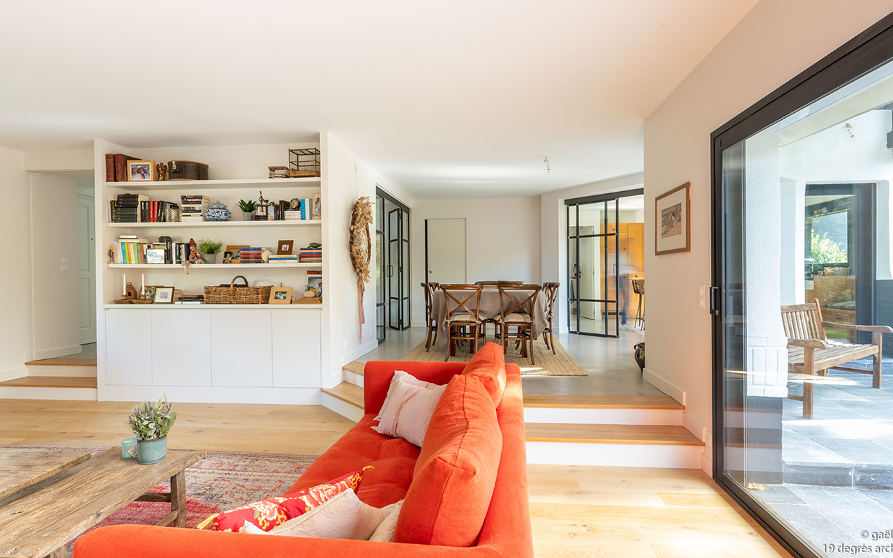 Salon avec un canapé orange, une table basse en bois brut et un agencement sur-mesure donnant sur la salle à manger. Le salon est sur un niveau inférieur, il y a 2 marches pour accéder à la salle à manger.