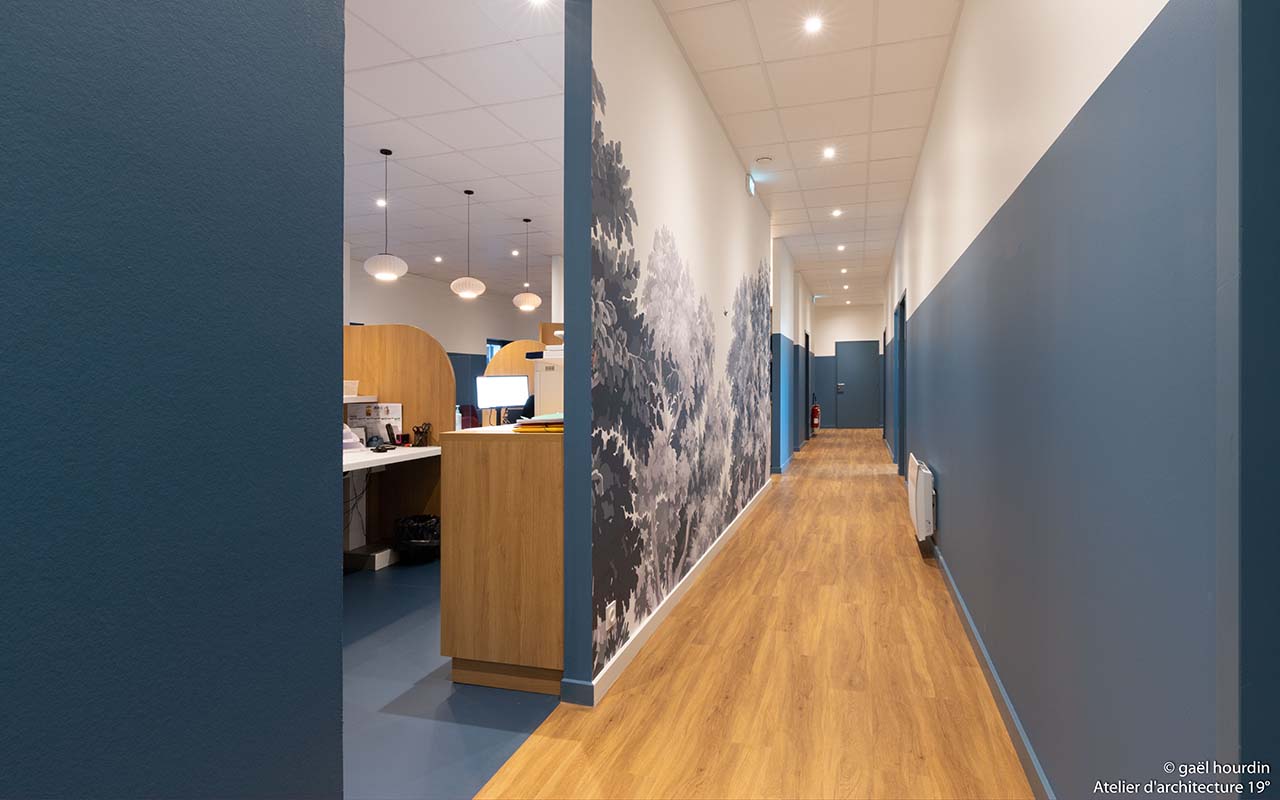Couloir menant aux différentes salles de soins. Une tapisserie florale blanche et noir et de la peinture bleue habillent le mur.