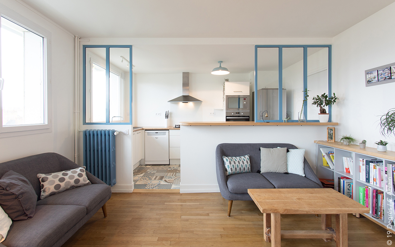 Salon avec deux canapé en tissu gris, une table basse en bois et un meuble à case bibliothèque. Un demi mur avec une verrière permet de dissocier l'espace du salon de la cuisine.