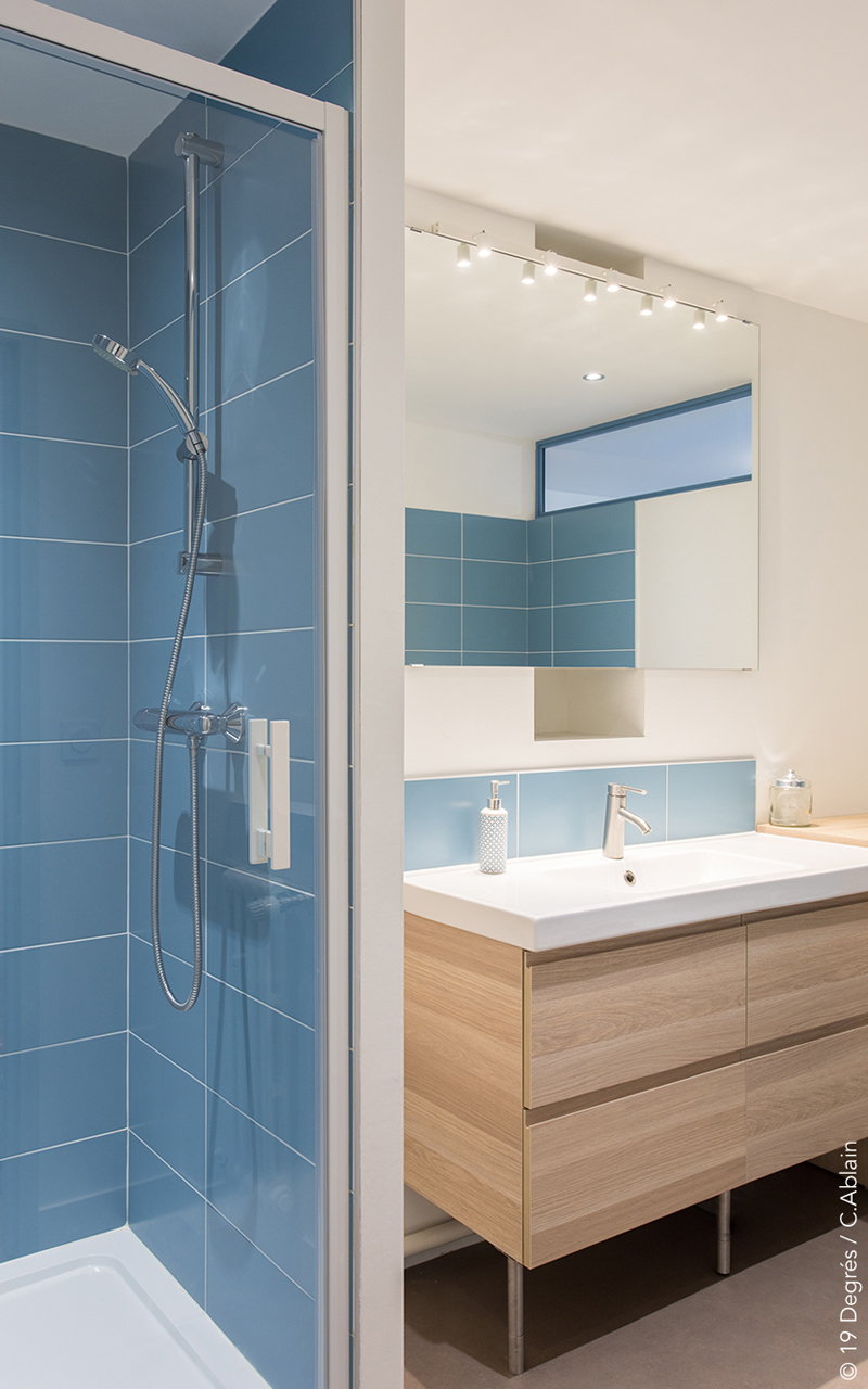 Salle de bain avec une douche carrelée bleu et un vasque blanc sur un meuble bois clair.