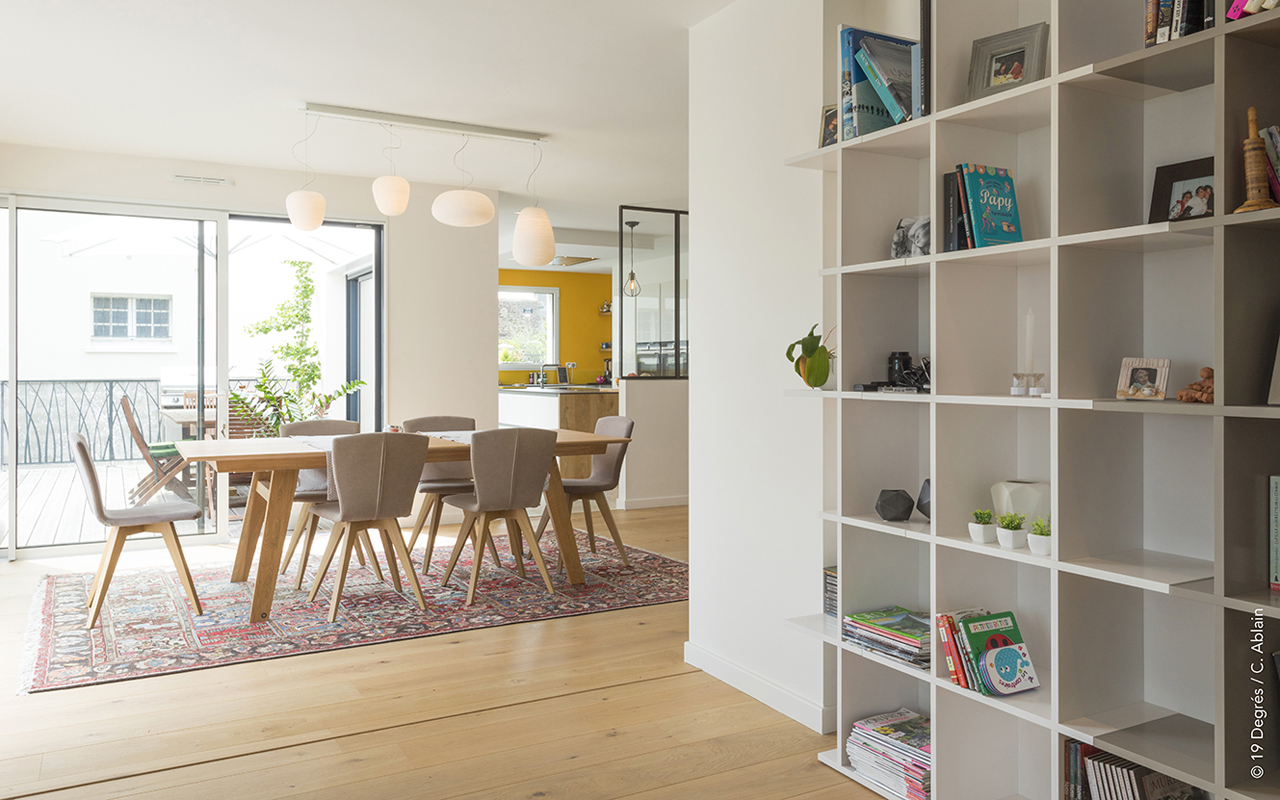 Salon avec table en bois et chaises type scandinave. Vue sur la cuisine et un meuble à case.