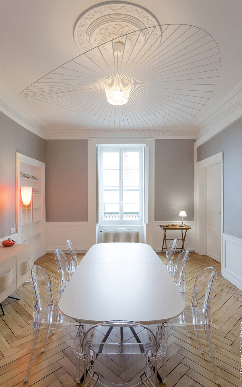 Salle à manger avec une grande table blanche, des chaises transparentes, un plancher en bois et un lustre blanc.