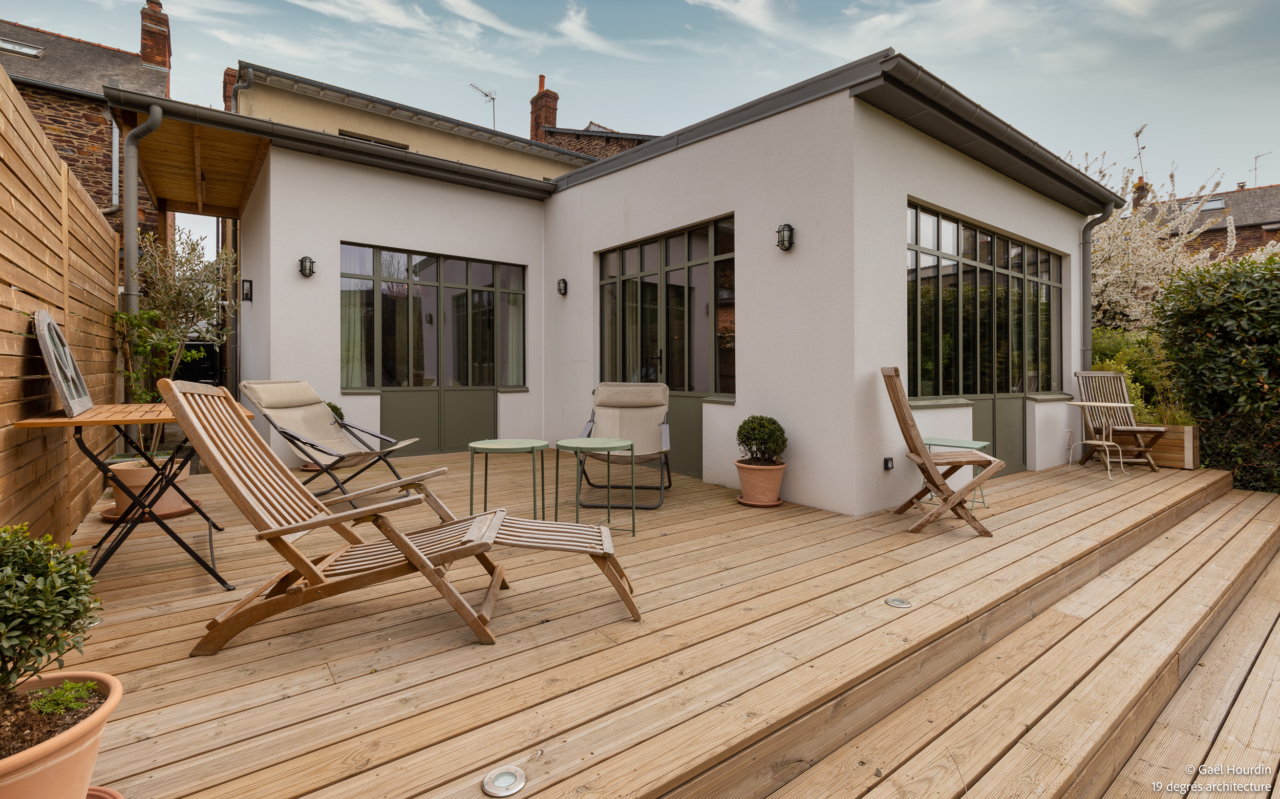 Extension dans son ensemble avec un enduit blanc et des verrières vertes. Une terrasse y est attenante en lame de bois. Du mobilier de jardin est présent sur cette terrasse.