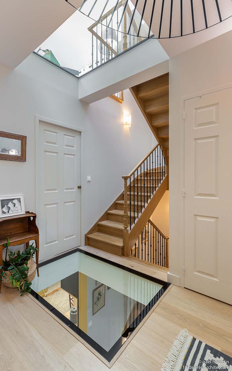 Etage avec vue sur l'escalier. Le sol comporte une verrière pour apporter de la luminosité. Les murs sont blancs et l'escalier est en bois.