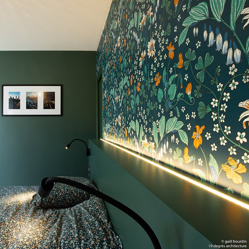 Détail d'une chambre. Lit double et murs sur les tons verts. Tapisserie florale au dessus de la tête de lit.