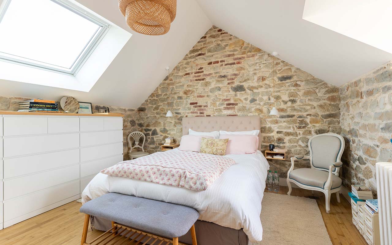 Chambre dans son ensemble avec des murs en pierres, un lit double et un meuble sur-mesure blanc et bois.