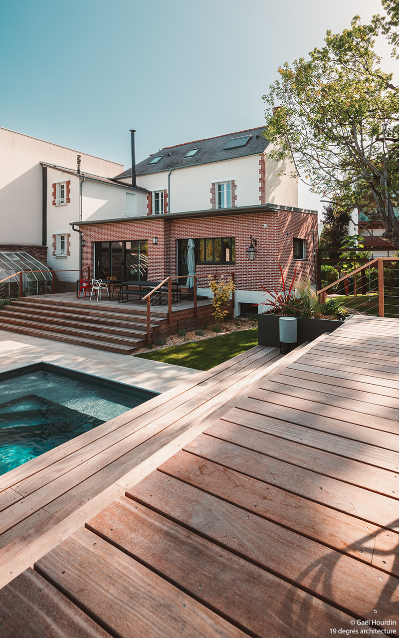 Maison blanche, extension en brique rouge et piscine. Vue de la terrasse en bois.