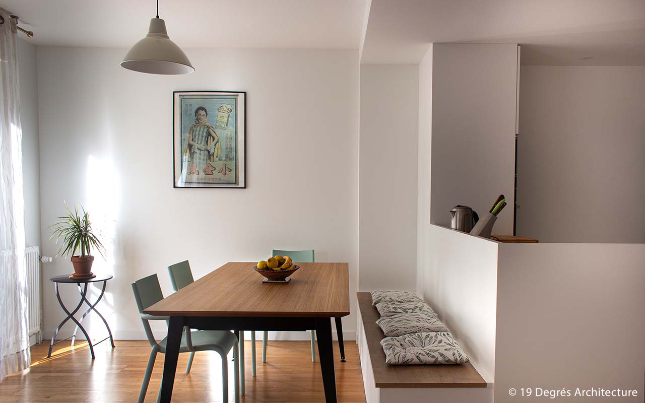 Salon cuisine d'un appartement. Les couleurs sont claires, on y retrouve du bois pour la table et le parquet, du blanc pour la cuisine et les murs et un cadre de plusieurs couleurs.