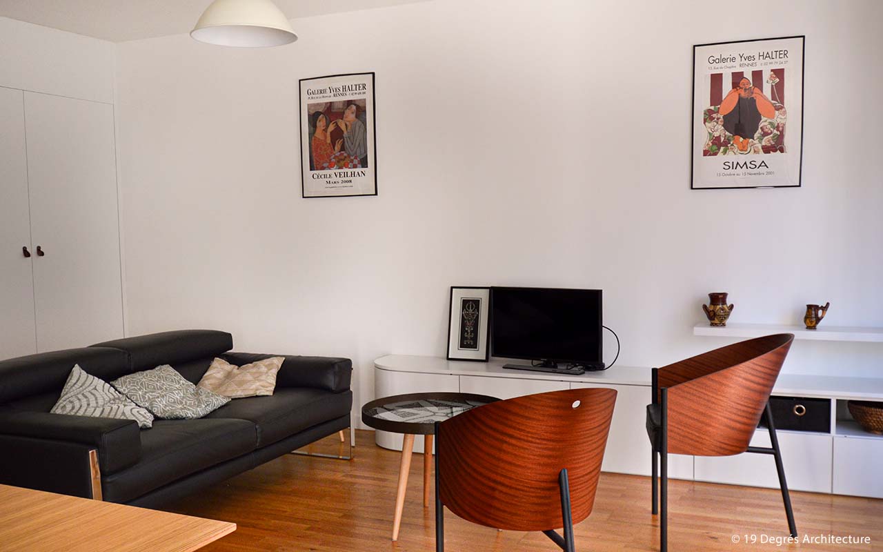 Salon présentant un canapé en cuir noir ainsi que des fauteuils en bois. On y voit également un meublé télévision blanc, une télévision et deux cadres accrochés sur un mur blanc.