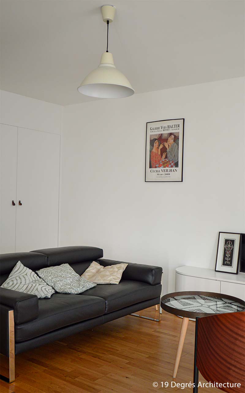 Salon d'un appartement présentant un canapé en cuir noir, un sol en parquet et un meuble télévision blanc. Il y a également un luminaire banc suspendu au plafond.
