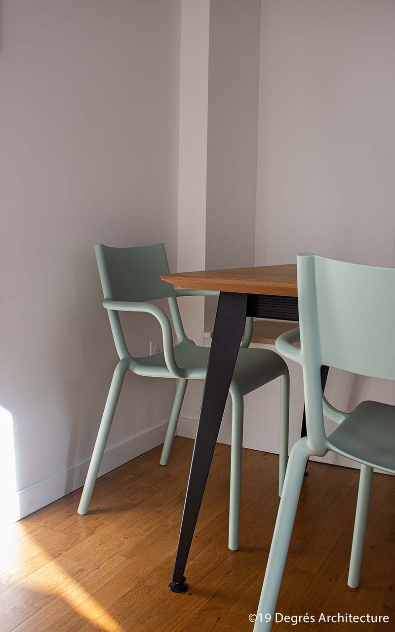 Détail du mobilier. Table en bois avec pieds en métal noir. Chaises en plastique dur de couleur vert pastel.