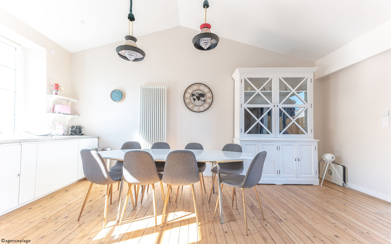 Cuisine salle à manger bord de mer dans des tons épurés. Le sol est en bois, la table et les chaises sont de type scandinave. Les meubles et la cuisine sont blancs.