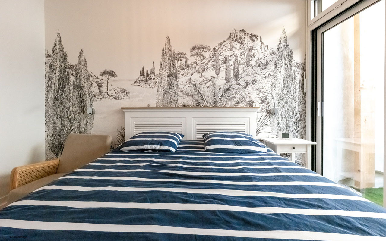 Chambre d'une maison bord de mer. Tapisserie noir et blanche présentant un paysage dessiné. draps rayés bleu marine et blanc et tête de lit blanche en bois.