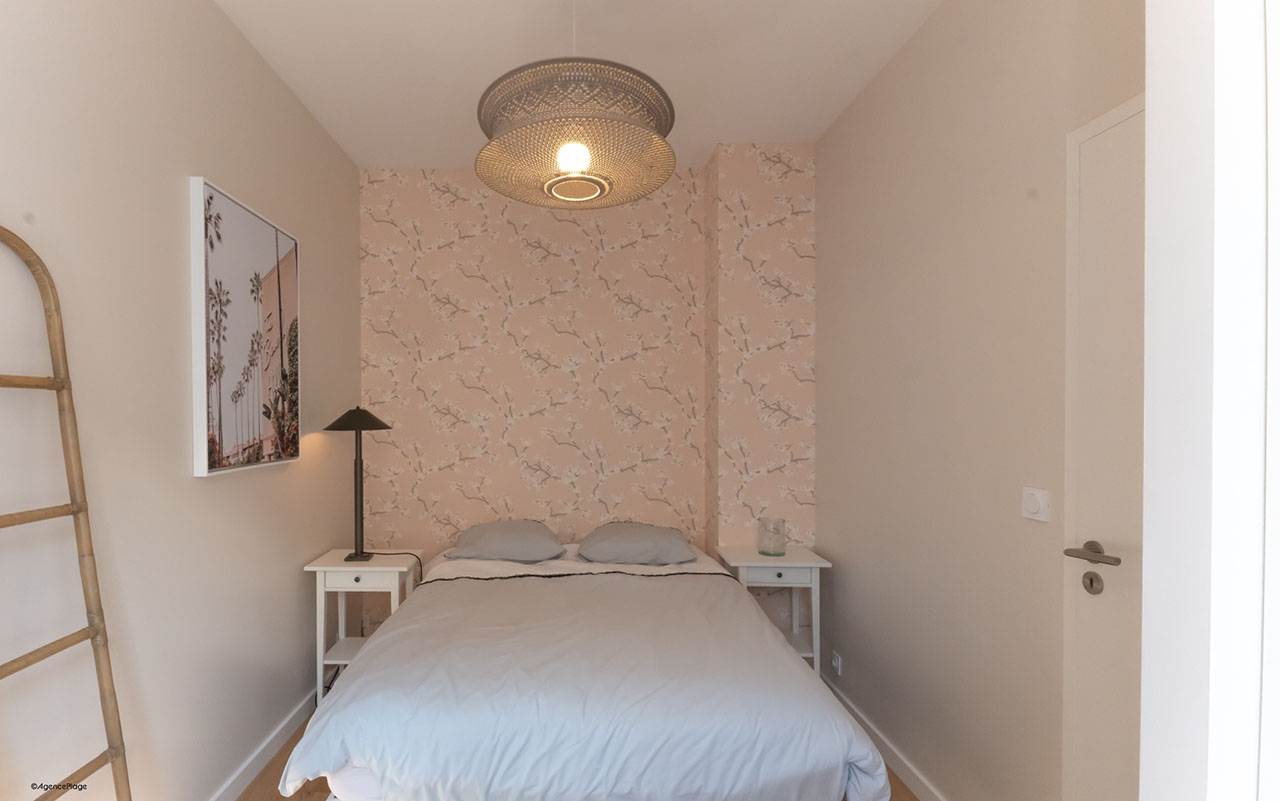 Chambre maison bord de mer avec un lit double, une tapisserie rose, un lustre et deux tables de chevet.