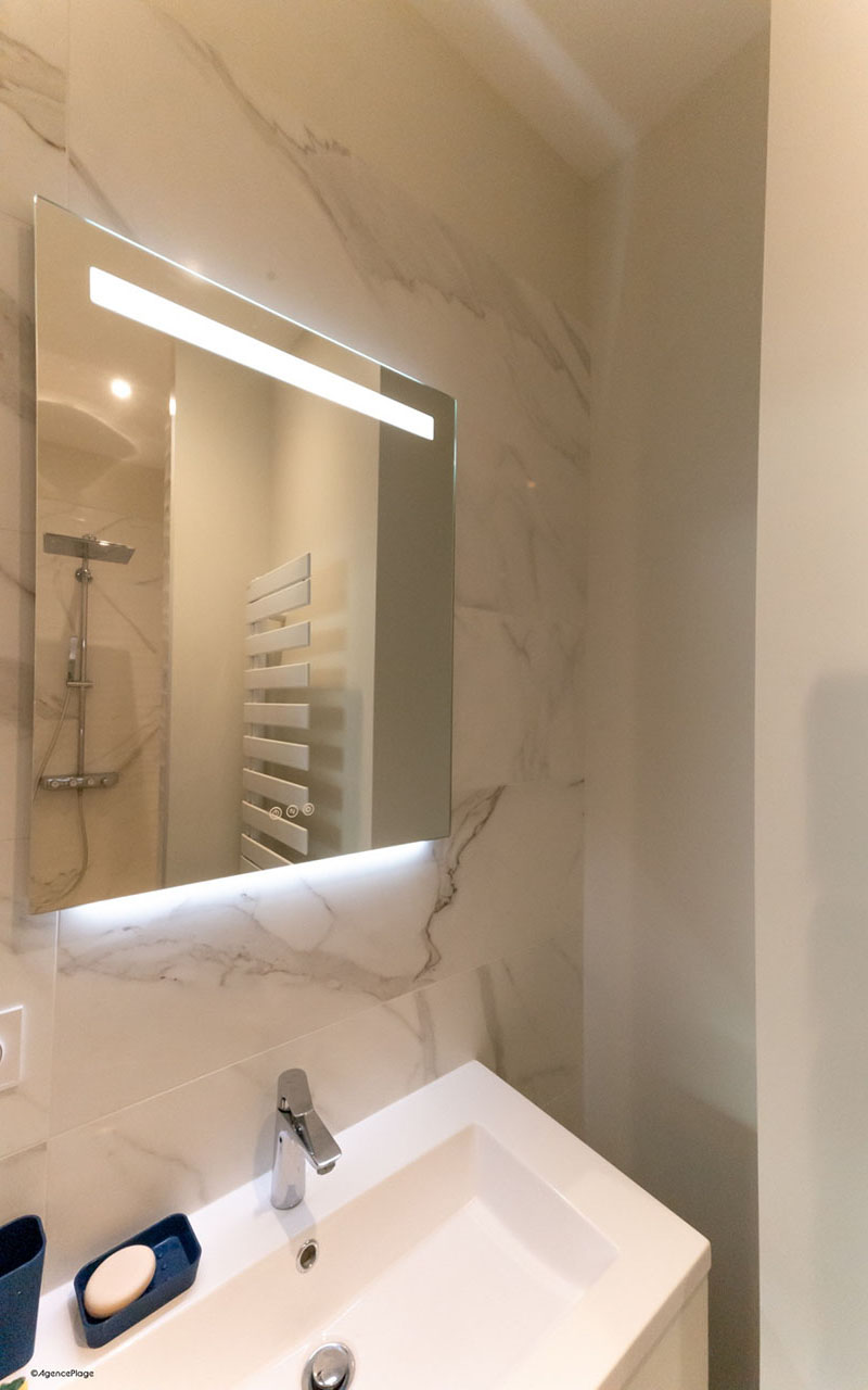 Salle d'au d'une maison en bord de mer. Les murs sont en carrelage effet marbre. Le vasque est blanc. La robinetterie de couleur argent et il y a un miroir au dessus du vasque.